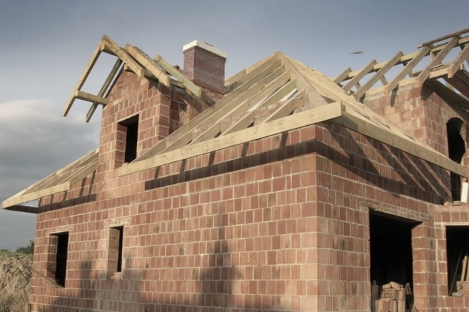 Surowy budynek w trakcie wznoszenia z cegły z widoczną konstrukcją dachu z belek drewnianych i kominem w ceglanej obudowie, otworami okiennymi i drzwiowymi bez ram